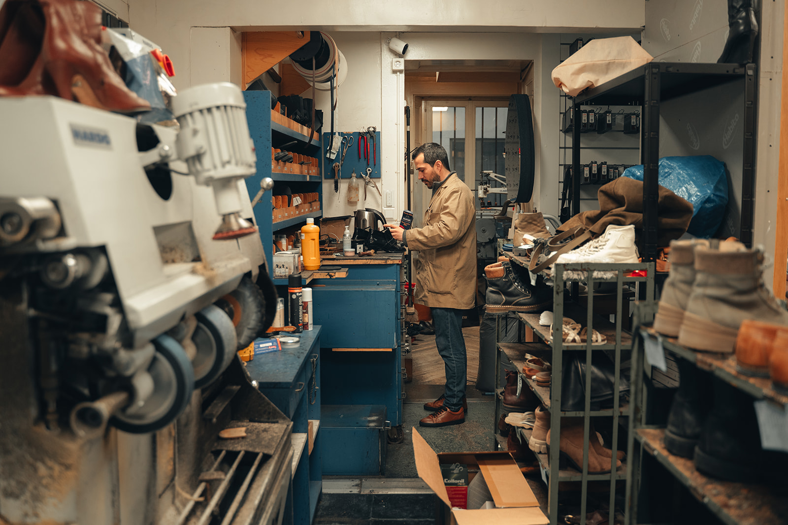 Cordonnerie de Neuchâtel 1 Artisan cordonnier dans son atelier à Neuchâtel, entouré d’étagères remplies de chaussures et de matériel de réparation.