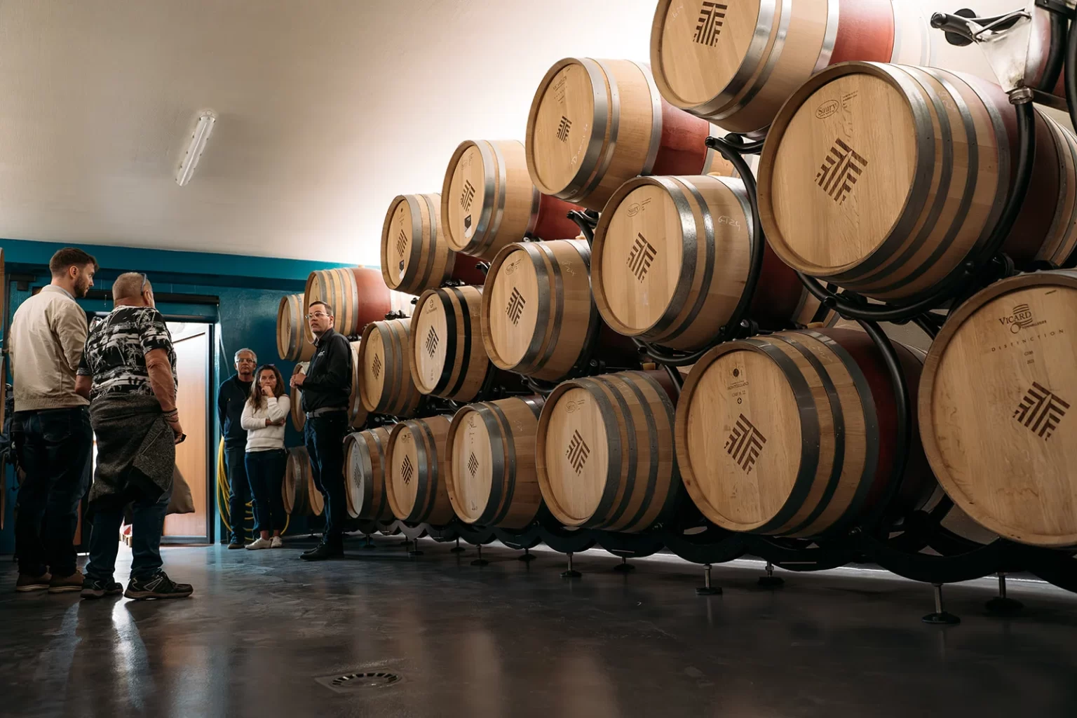 Alignement de barriques en bois dans une cave à vin moderne, avec visiteurs en arrière-plan.