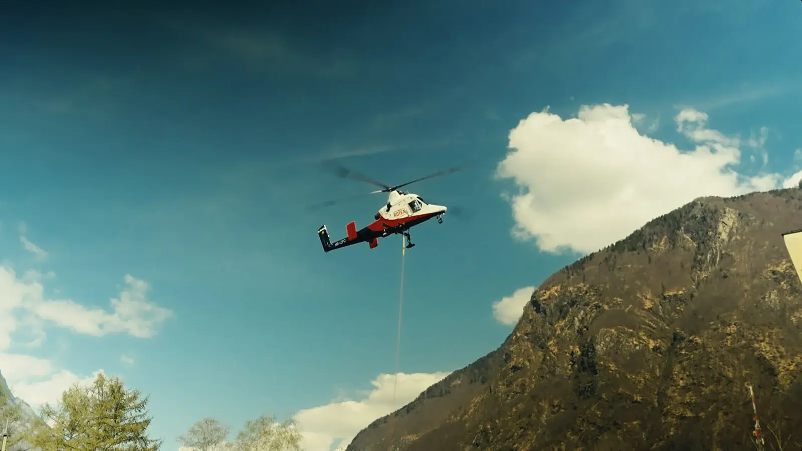 Hélicoptère en vol transportant une bobine de câble dans une vallée alpine après les inondations.